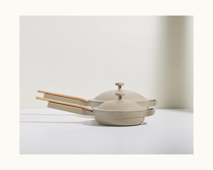 Two beige Mini Always Pan 2.0 stovetop pans with matching lids and light wood handles are stacked on a white surface, forming a stylish and convenient kitchen duo against a plain backdrop.
