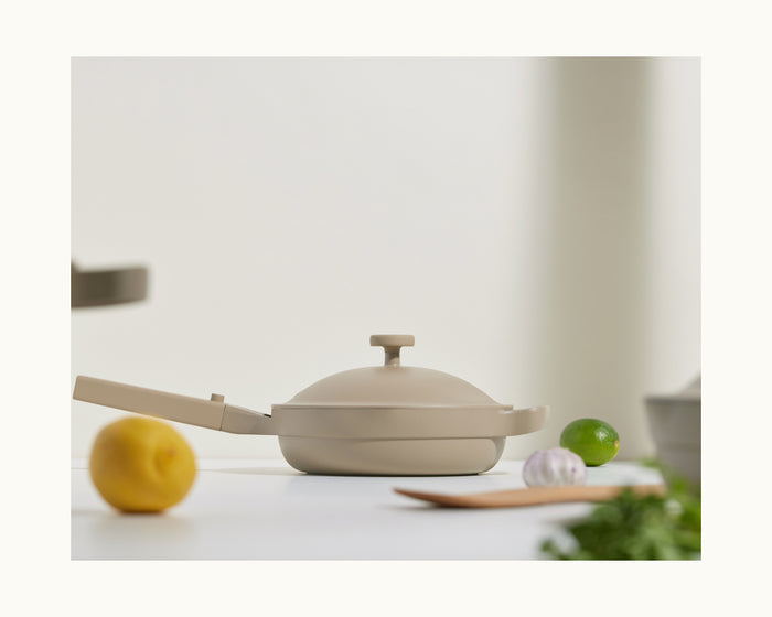 The Mini Always Pan 2.0, shown with a beige lid and handle, sits on a white surface among a lemon, lime, garlic, wooden spatula, and fresh herbs—perfect for stylish and versatile cooking in any kitchen.