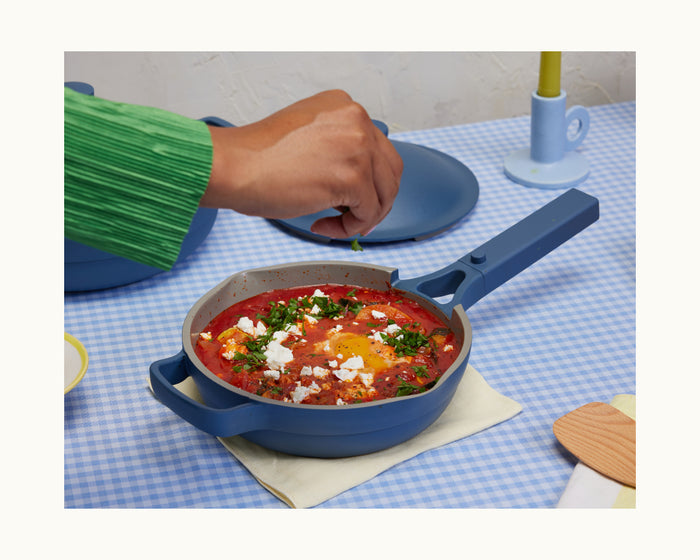 A person sprinkles herbs over a tasty tomato, egg, and cheese dish cooked in the Mini Always Pan 2.0, creating a convenient meal served on a blue checkered tablecloth.