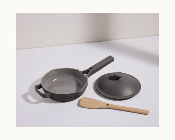 The Mini Always Pan 2.0, a stylish black nonstick frying pan with a matching lid and light wooden spatula, is arranged on a white surface for easy and convenient cooking in any kitchen.