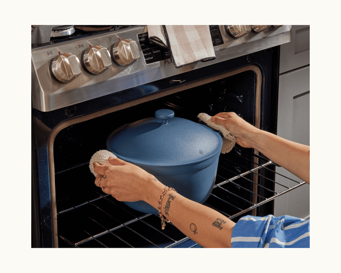 Using pot holders, a person puts the Perfect Pot, a family-sized 8-in-1 nonstick pot, into an oven. Stove knobs and part of the kitchen are visible in the background.