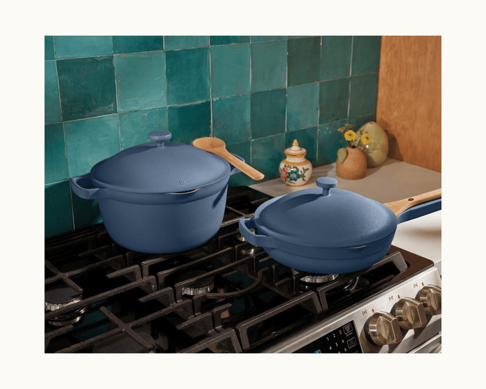 The Perfect Pot and a matching blue pan with lids and wooden handles sit on a gas stove in a kitchen featuring green tile backsplash and small countertop decor.