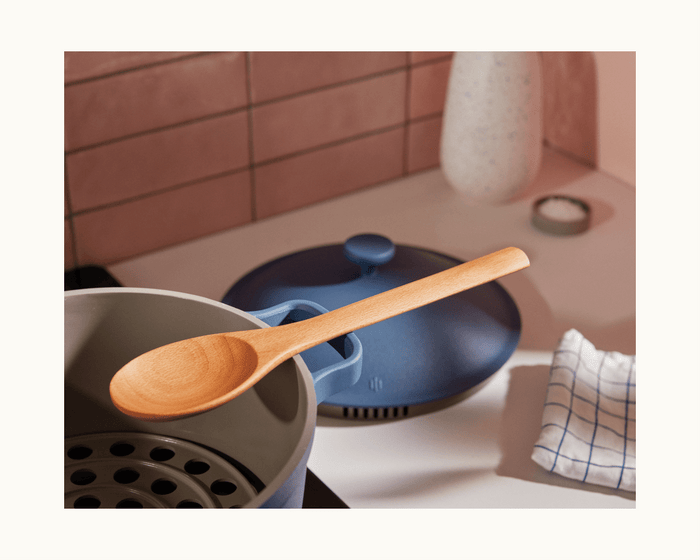 A wooden spoon rests on the edge of the family-sized Perfect Pot in blue, with its lid nearby; a checkered cloth and a vase are on the countertop in the background.