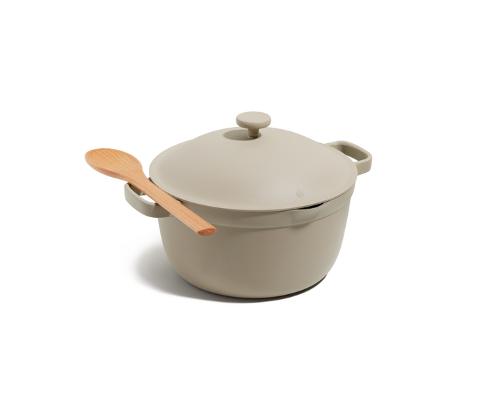 The Perfect Pot, a beige Dutch oven with two handles and a lid, is shown with a light wooden spoon on top—this versatile family-sized pot is ideal for cooking hearty meals.