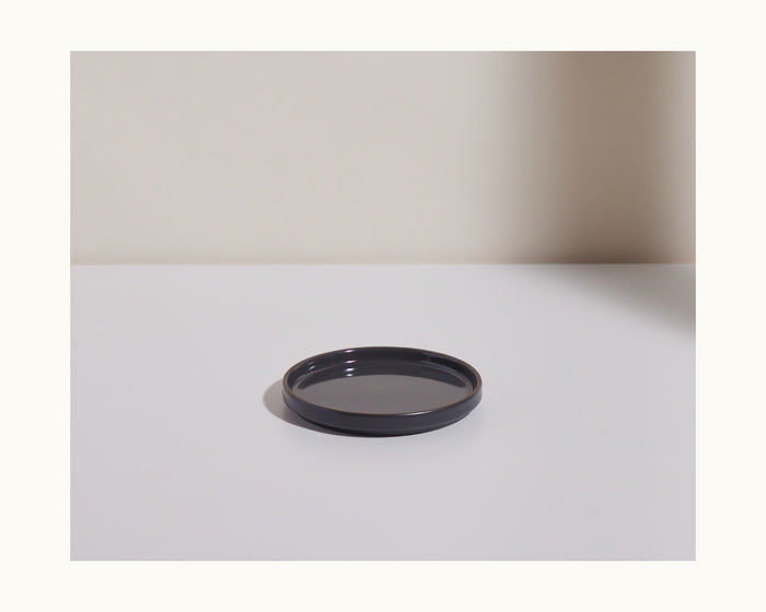 A round, shallow black ceramic Dessert Plate rests on a flat white surface against a light-colored background, highlighting its scratch-resistant finish.
