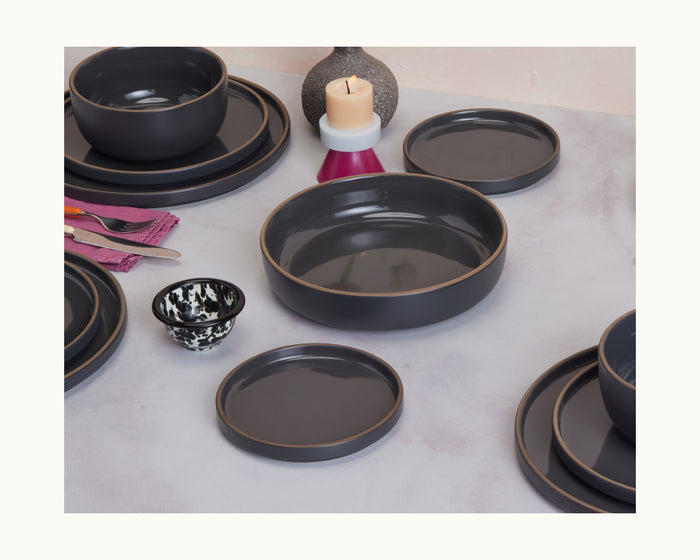 A table set with Dessert Plates, scratch-resistant ceramic plate, dark gray bowls, black and white cup, stacked plates, a candle, vase, cutlery, and a purple napkin completes this elegant tableware arrangement.