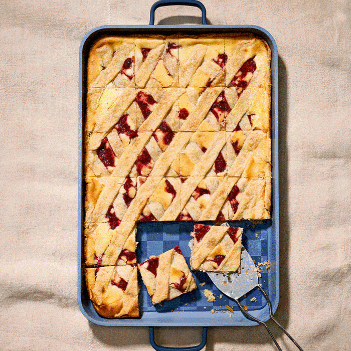 A rectangular Dinner at Our Place baking pan holds a lattice-topped jam bar dessert, with one square piece on a spatula—perfect for recreating Shiza Shahid’s latest cookbook recipes.