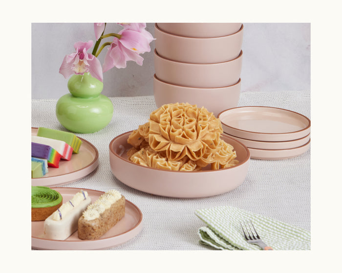 A table setting featuring pink dinner bowls with assorted desserts, a green vase filled with pink flowers, and a folded green checkered napkin holding a fork and spoon.
