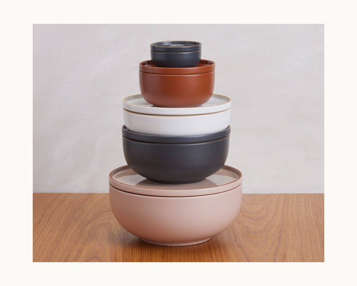 A stack of five colorful Dinner Bowls in assorted sizes sits on a wooden surface, showcasing a vibrant tableware set against a light background.