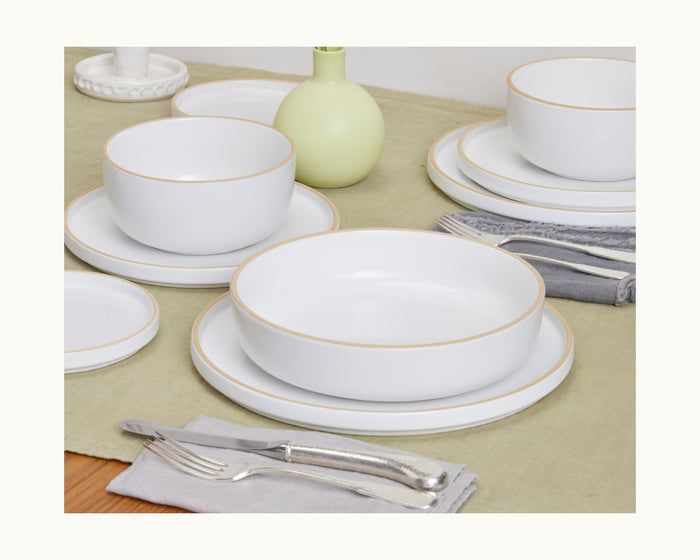 Dinner Bowls with gold rims and a matching shallow plate are displayed on a light green tablecloth, accompanied by silverware, napkins, and a small green vase.