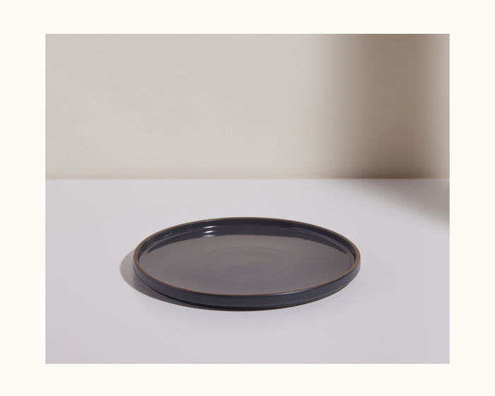 A dark gray hand-glazed Dinner Plate rests on a white surface with a beige background, ideal for enhancing your tableware collection.