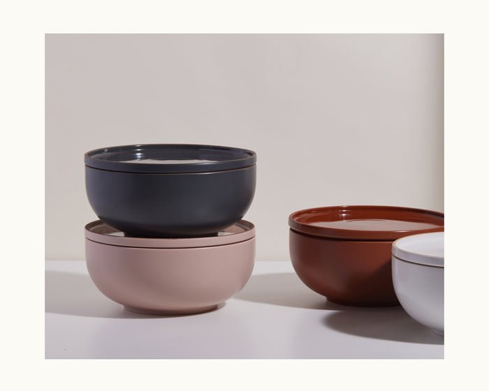 Four dinner plates in black, pink, brown, and white are stacked on a white surface against a neutral background, bringing elegance to your tableware collection.