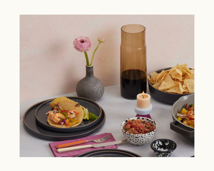 A table set with tacos, tortilla chips, salsa, a lit candle, two vases—one tall and amber, one small with a pink flower—Dinner Plates, utensils, all against a light pink background.