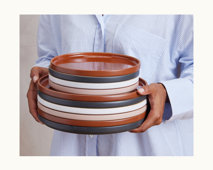 A person in a light blue striped shirt holds a stack of Dinner Plates—hand-glazed round ceramic tableware with brown, white, and black stripes.