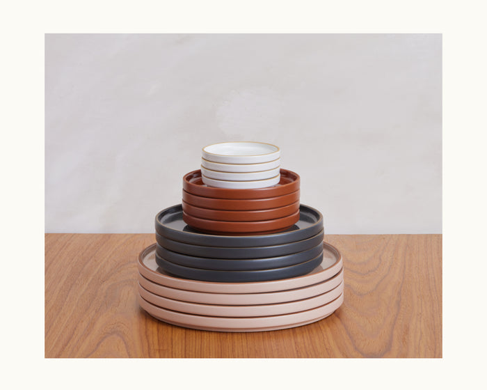 A stack of hand-glazed Dinner Plates in beige, dark gray, and rust rests on a wooden surface against a light wall, highlighting artisanal tableware.
