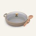 The Dual Handle Always Pan, a beige and versatile nonstick pan featuring a glass lid with a gold knob, sits next to a wooden spoon on a plain light background.