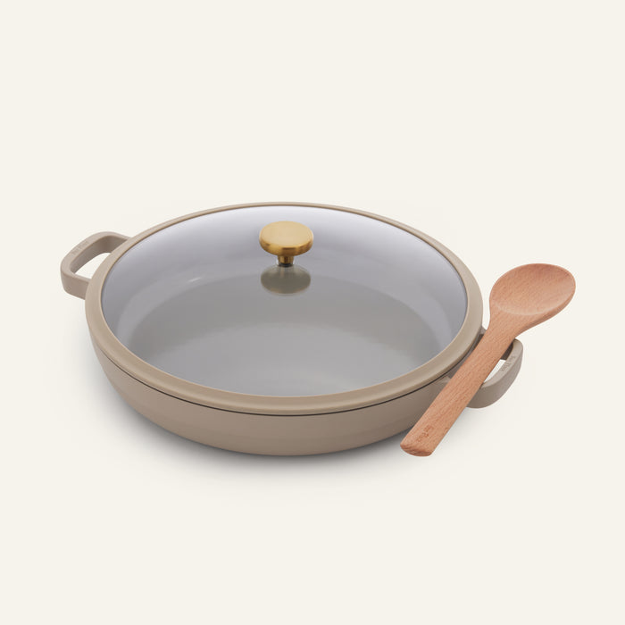 The Dual Handle Always Pan, a beige and versatile nonstick pan featuring a glass lid with a gold knob, sits next to a wooden spoon on a plain light background.