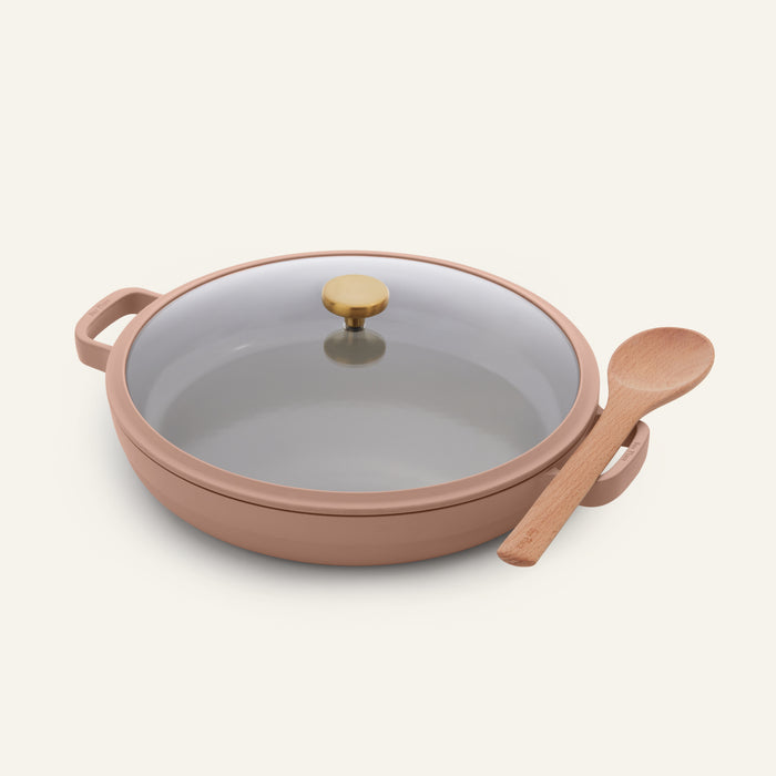 The Dual Handle Always Pan is a versatile, beige nonstick pan featuring a glass lid with a gold knob, shown with a wooden spoon resting on its handle against a plain light background. This non-toxic cookware is perfect for everyday use.