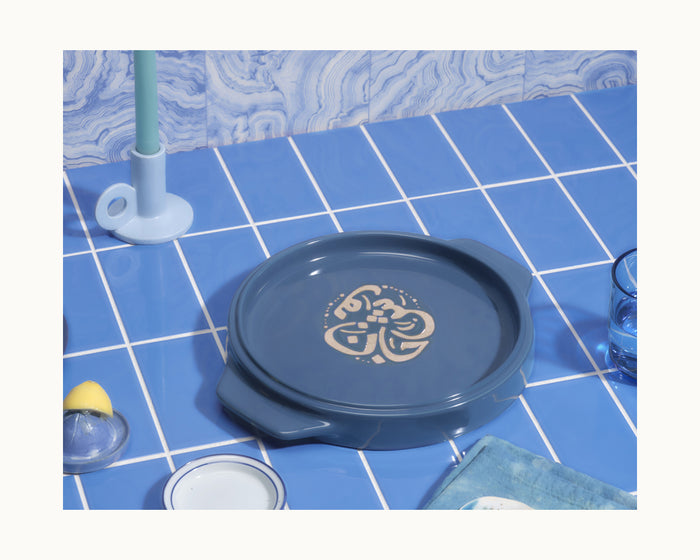 The Flipping Platter, a round blue baking dish with beige accents, sits on a tiled countertop amid blue tableware and a candle—ideal for serving maqluba or tahdig and perfect as an elegant flipping platter.