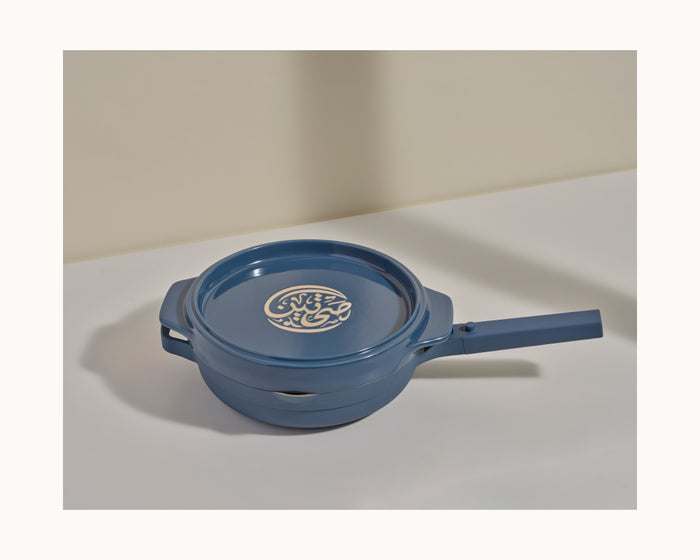 The Flipping Platter, featuring a blue lid with a long handle and decorative design—perfect for making tahdig—rests elegantly on a white surface with a light beige backdrop.