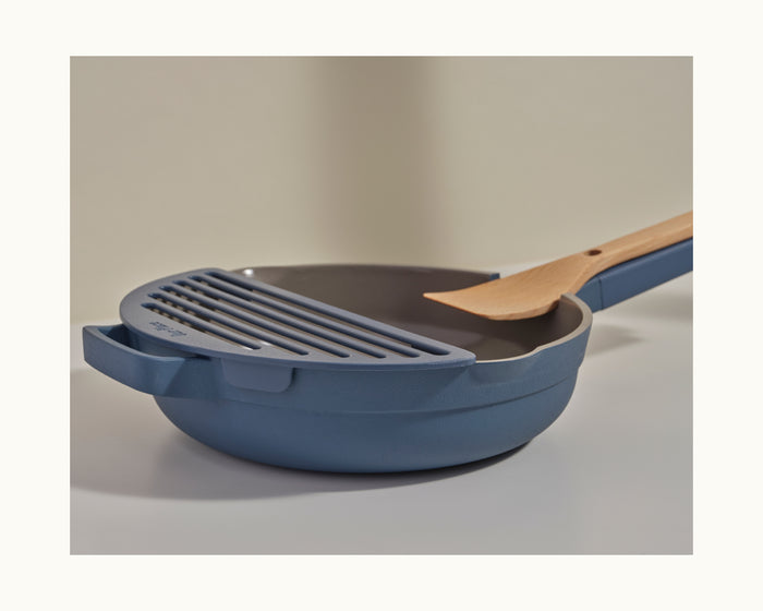 A blue nonstick frying pan from Our Place, paired with a wooden spatula and the matching blue Fry Deck accessory, displayed on a neutral surface.