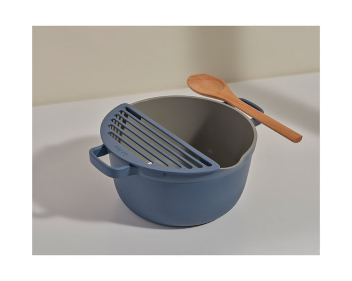 The Fry Deck from Our Place is a blue cooking pot with a built-in strainer lid and wooden spoon, resting on a white surface—a versatile addition to any frying accessory collection.
