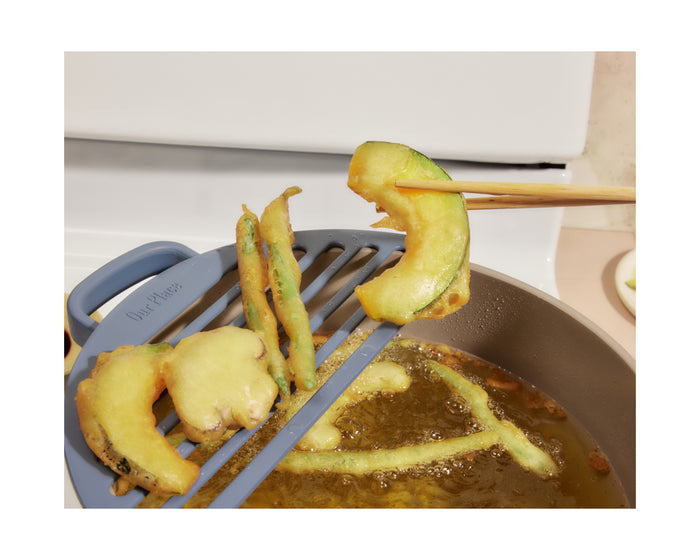 A hand uses chopsticks to place fried vegetable tempura on the blue Our Place Fry Deck, which sits above a pot of hot oil on the stove.