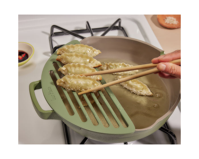A hand uses chopsticks to pick up fried dumplings from a pan with the Fry Deck draining rack, highlighting this convenient Our Place cookware accessory on the stovetop.