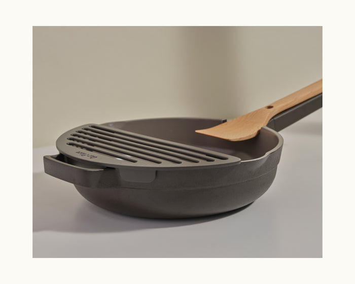 A Fry Deck slotted metal lid from Our Place rests partially atop a black nonstick pan with a wooden-handled spatula inside, all set on a white surface.