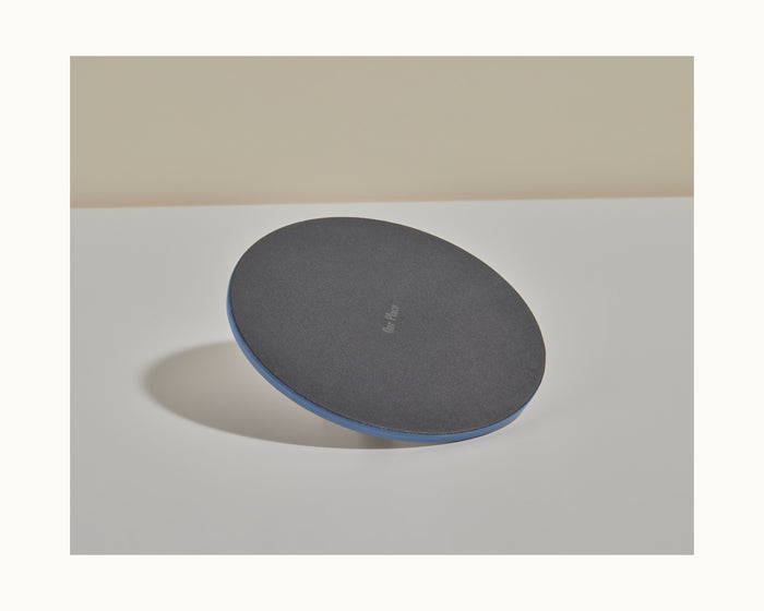 A black, round Grill Press with a blue edge sits on a white surface, casting a shadow to the left. Known for even heat distribution, this cast iron object aids in efficient grilling.