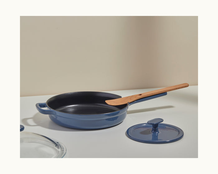 A blue Grill Press with a matching lid and a wooden spatula on its handle, set on a white surface against a neutral background, ensures even heat distribution for perfect grilling every time.