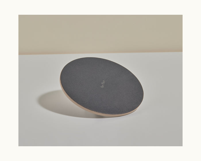 A round, flat Grill Press with a dark cast iron-like surface and a light-colored edge sits on a light gray surface against a beige background, designed for even heat distribution.