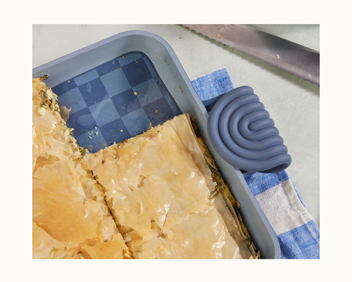 A rectangular baking tray with Hot Grips helper handles sits on a blue and white towel next to a serrated knife, providing extra kitchen protection.