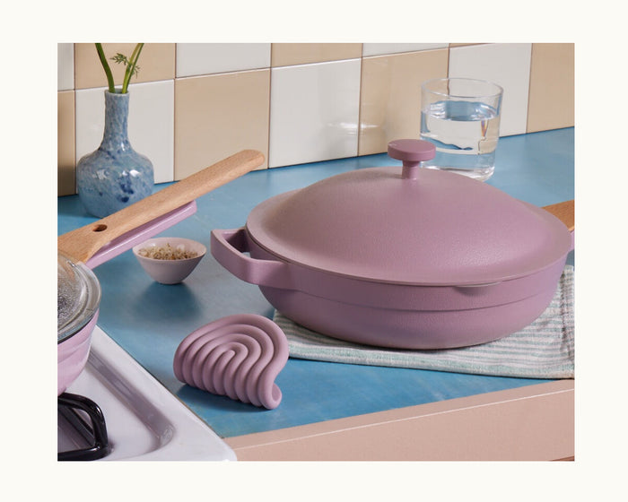 A pink cooking pot with helper handles and a lid sits on a blue countertop beside Hot Grips, a glass of water, a small bowl of grains, and a vase with green stems.