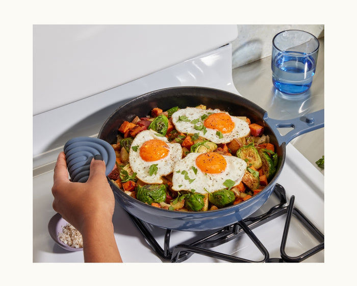 A hand holds a skillet with fried eggs, Brussels sprouts, and veggies on the stove using Hot Grips for kitchen safety, beside a glass of water and a small bowl of salt.