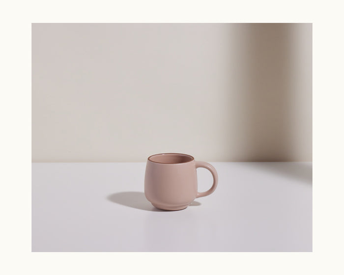 A Night + Day Mug in plain pink ceramic stoneware rests on a white surface, with a soft shadow cast to the left, set against a light beige background.