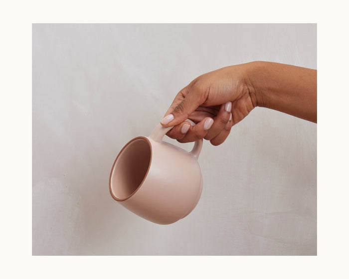 A hand with light nail polish holds the light pink Night + Day Mug by the handle against a plain, light background.