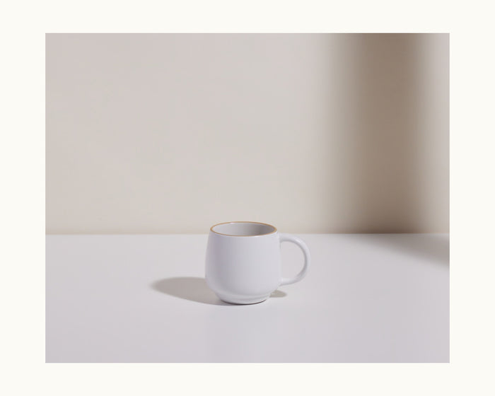 The Night + Day Mug, a plain white ceramic stoneware cup, sits on a white surface with a soft shadow against a neutral background.