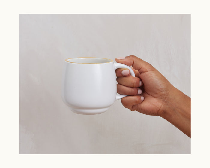 A hand holds a Night + Day Mug, a plain white ceramic stoneware cup, against a neutral background.
