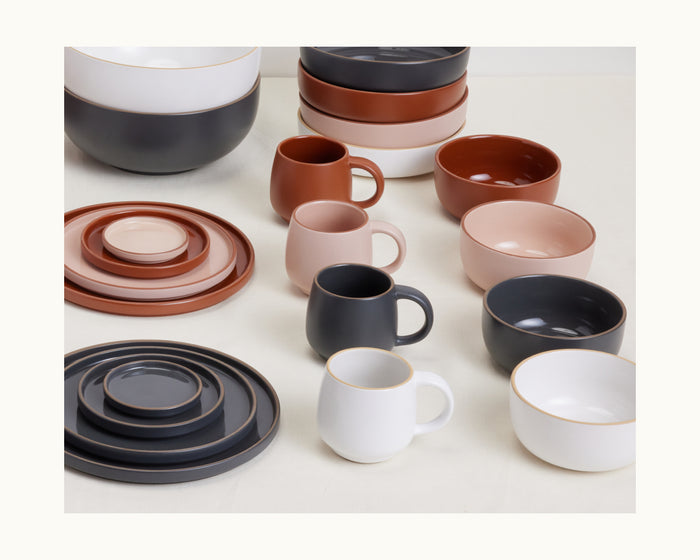 Night + Day Mugs in shades of white, terracotta, pink, and dark gray are displayed alongside ceramic plates and bowls on a light surface.