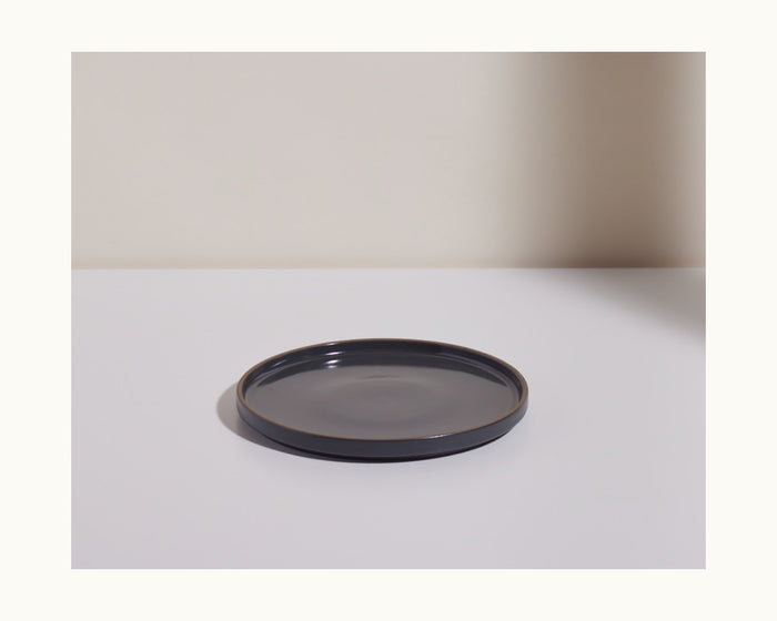 A Salad Plate in dark gray with a hand-glazed finish rests on a white surface, set against a light-colored wall.