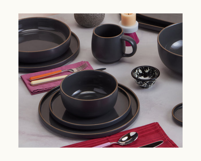 Salad Plates in matte black are arranged with bowls, mugs, silverware, small decorative bowls, and folded napkins on a light surface.