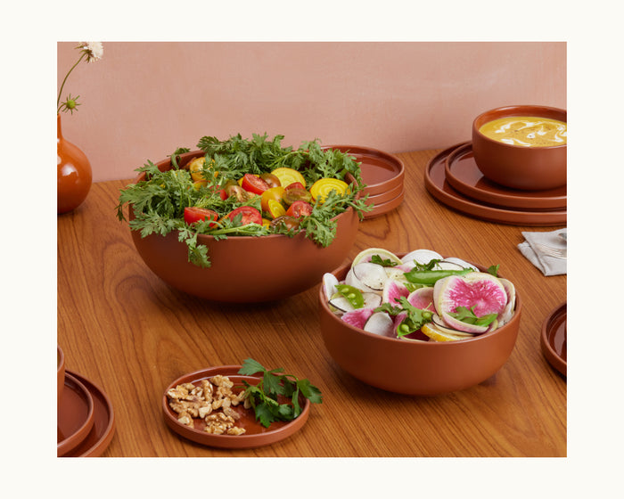 Two Serving Bowls rest on a wooden table—one filled with mixed greens and tomatoes, the other with sliced radishes and onions—while walnuts and parsley nearby complete this inviting tableware set, with soup in the background.