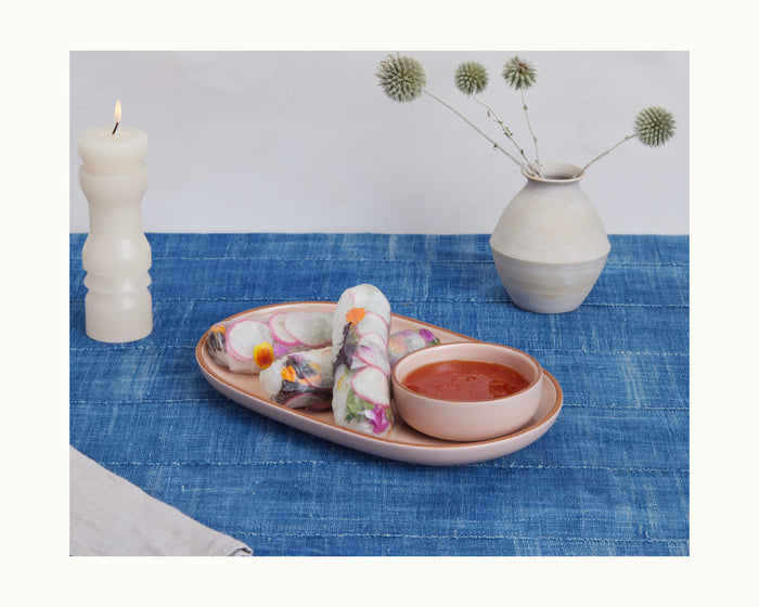 Two vegetable spring rolls on a pink Serving Platter with dipping sauce, set on a blue tablecloth beside a candle and thistle flowers. The perfect way to showcase appetizers and elevate your table setting with our Serving Platters.
