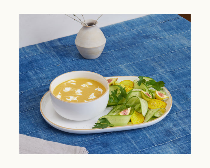 A ceramic Serving Platter holds a creamy yellow soup with white swirls next to a fresh salad of cucumbers, yellow tomatoes, figs, and parsley atop a blue cloth, complemented by elegant tableware and a vase in the background.