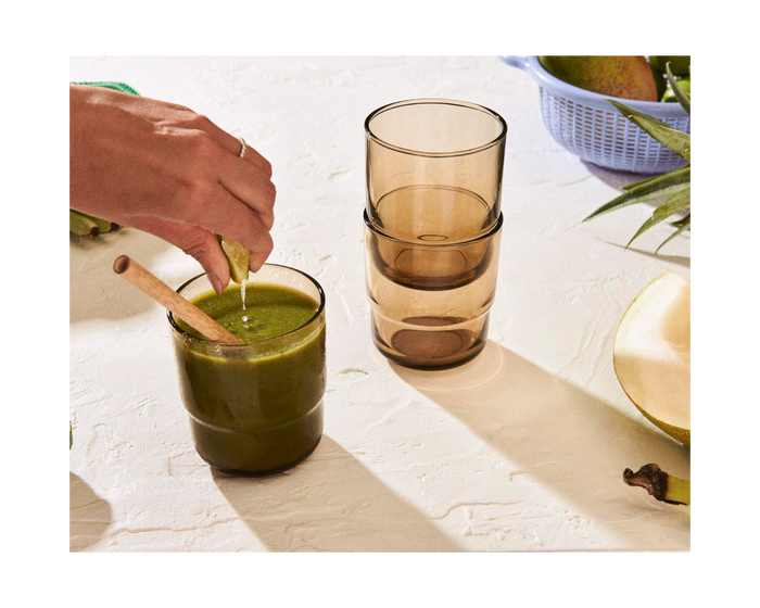 A person stirs a green drink in a glass with a wooden spoon; colorful drinkware, fresh fruits, and two stacked Short Night + Day Glasses are visible in the background.