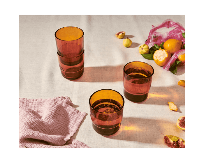 Four handmade Short Night + Day Glasses in amber sit on a light tablecloth, with a pink towel, scattered fruit, and a pink bag nearby—perfect for adding colorful drinkware to your table.