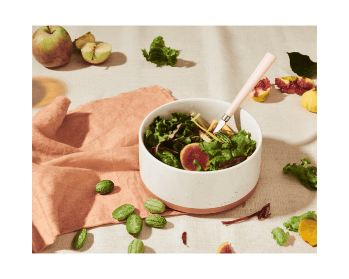 A hand-painted Side Bowl holds a mixed green salad with figs, placed on a beige tablecloth with a pink-handled fork, surrounded by sliced apple, leafy greens, and small green vegetables.