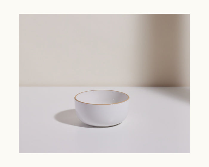 A Soup Bowl, small and white with a thin gold rim, from a charming ceramic set, sits on a plain white surface against a neutral background.