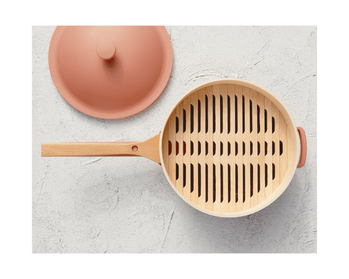 A round beige Spruce Steamer with a wooden handle and slotted wooden insert sits beside its matching lid on a light textured surface—perfect for pairing with bamboo cooking chopsticks.